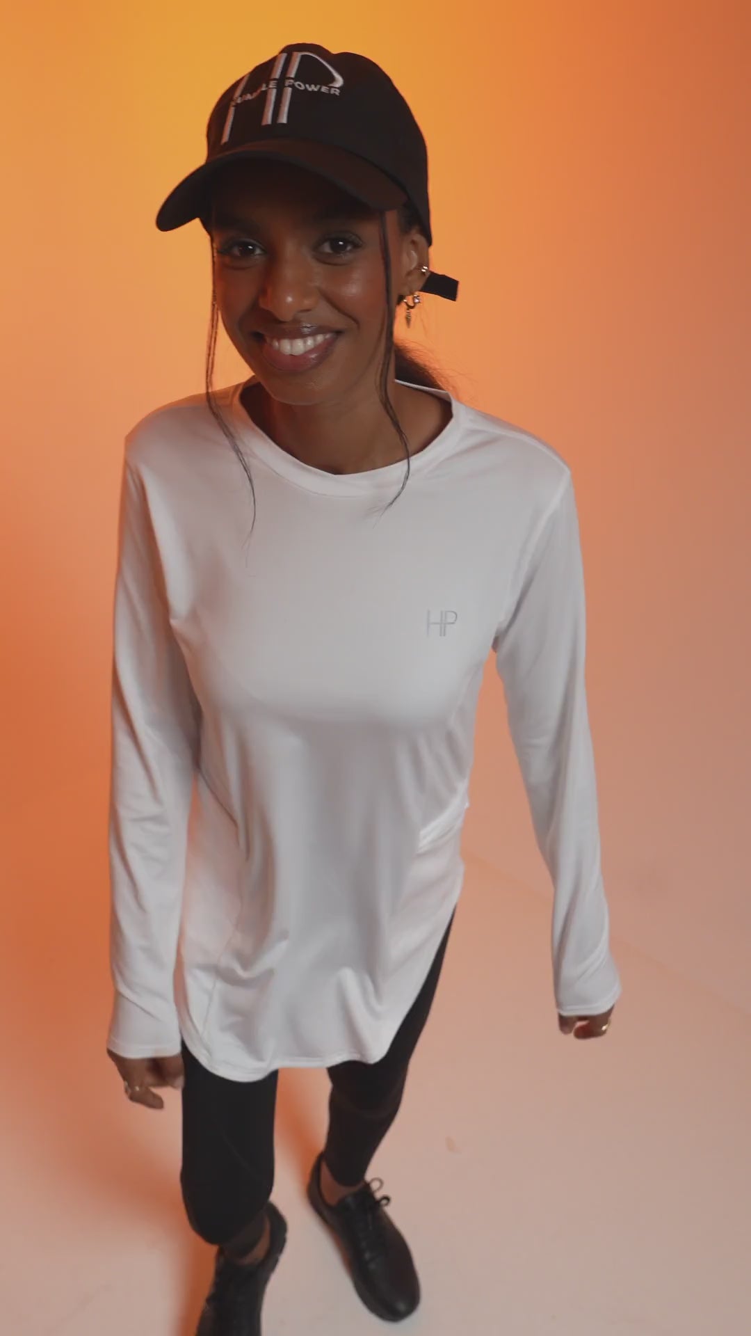 Woman in a white long-sleeve modest top and Humble Power cap on an orange background